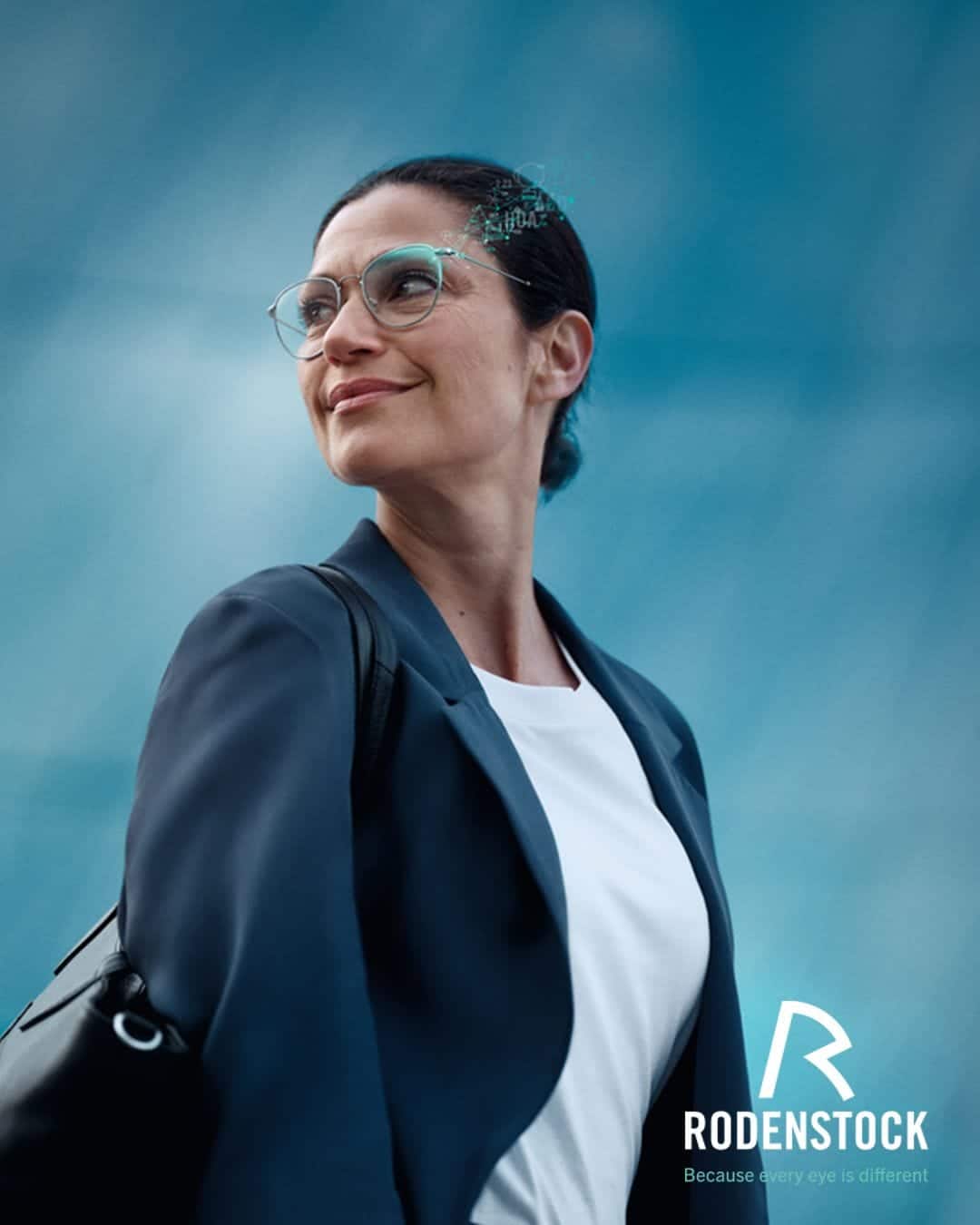 Woman wearing glasses and a blazer looks upwards with a slight smile, showcasing the clarity provided by Rodenstock lenses. The Rodenstock logo and slogan “Because every eye is different” are visible in the lower right corner.