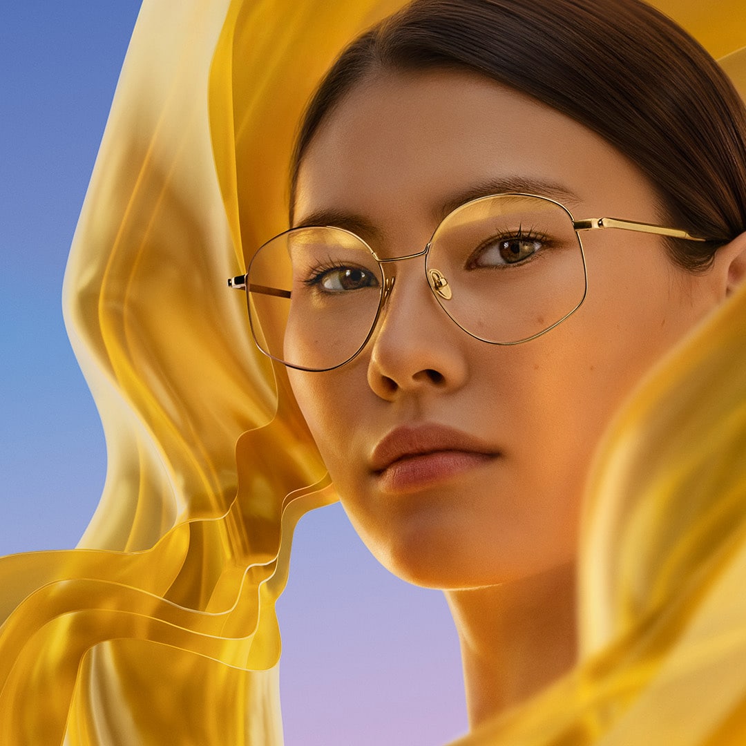 Woman wearing round gold glasses and looking forward, with a yellow, translucent fabric swirling around her face and a purple-blue gradient background.