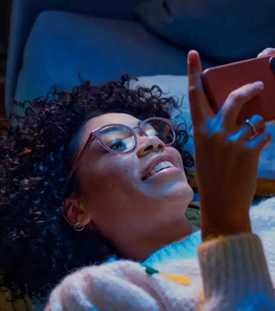 A person with curly hair and glasses lies on a couch, smiling while holding and looking at a smartphone, their clear vision thanks to Essilor quality lenses.