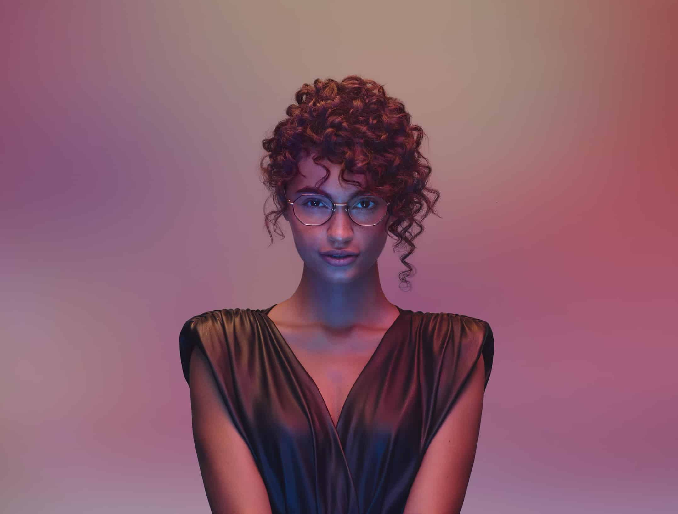 A person with curly hair and glasses, featuring high-quality Essilor lenses, wears a sleeveless, dark v-neck top, standing against a gradient pink and purple background.