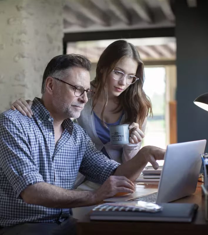 A man and a woman look at a laptop together at a desk. The man is typing while the woman, holding a mug, stands beside him. Both wear Essilor Lens glasses and appear focused on the screen.