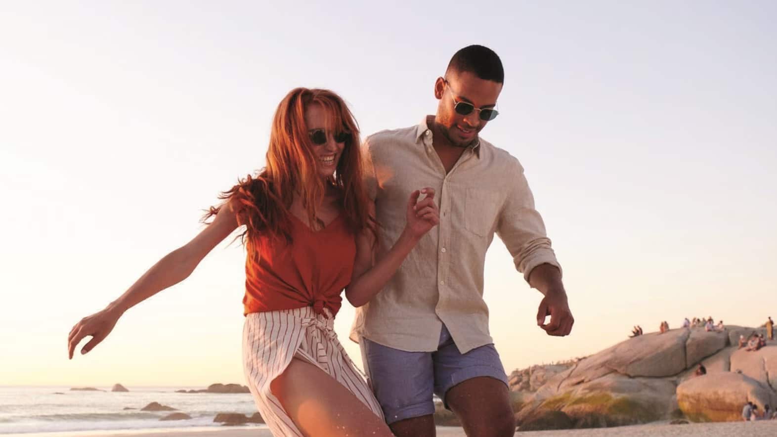 A woman and man in summer clothes walk barefoot on a beach, smiling and holding hands, with rocks and a clear sky in the background—a perfect moment captured through a quality lens.