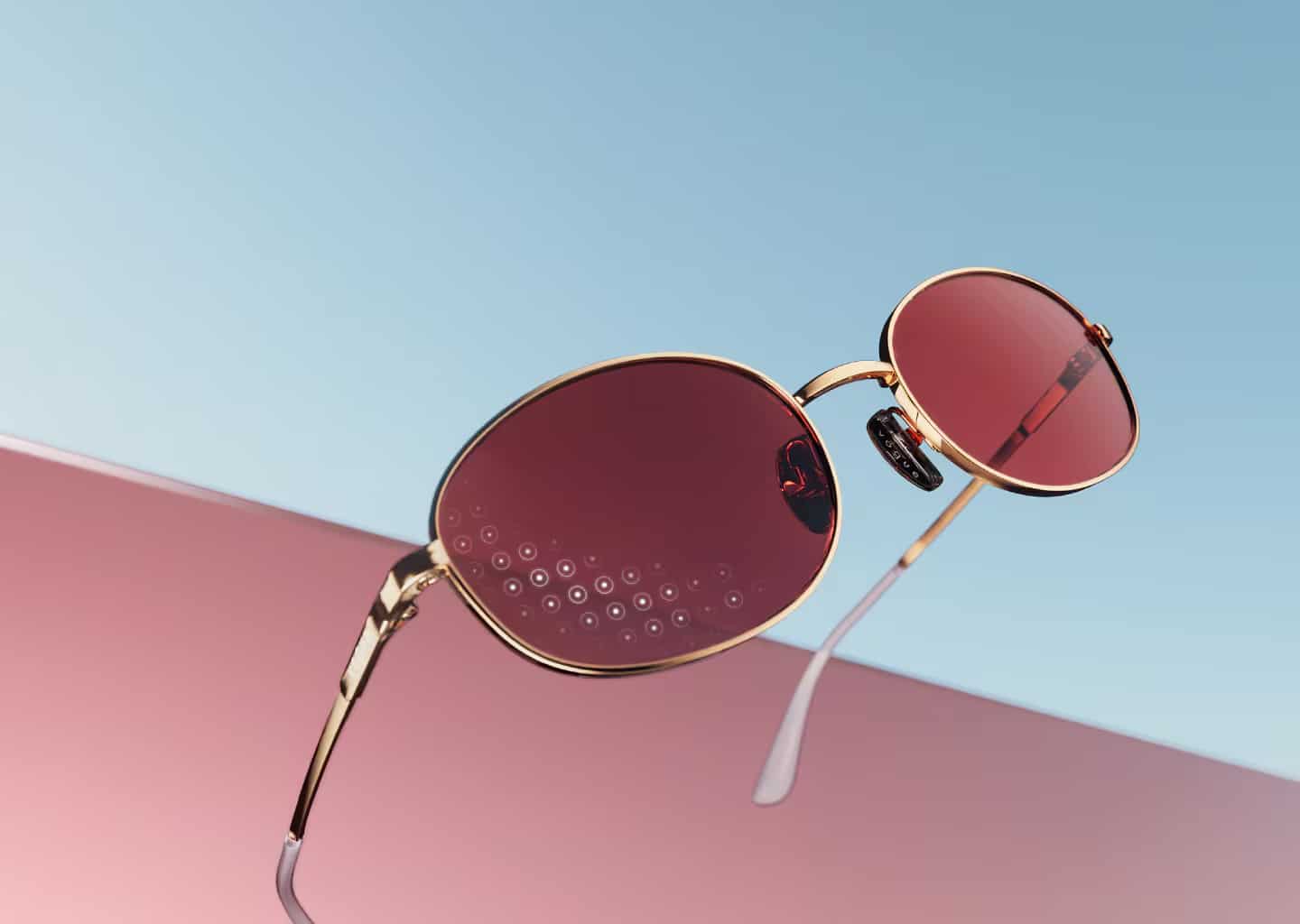 Gold-framed sunglasses with round red lenses rest on a pink surface against a blue background; one lens, featuring decorative dots, showcases Essilor quality lens craftsmanship.