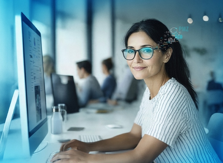 Woman with glasses featuring Rodenstock Lens working at a computer in a modern office, looking at the camera and smiling, with colleagues in the background.
