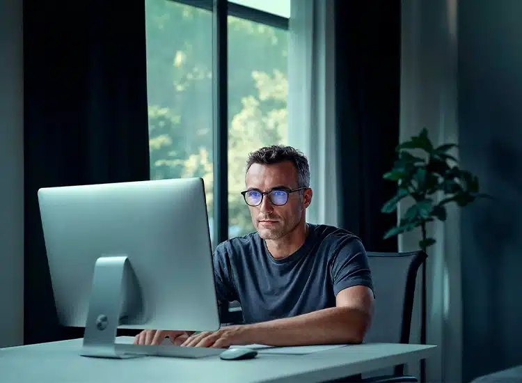 A man wearing glasses with Rodenstock eyeglass lenses sits at a desk working on a desktop computer in a modern room with large windows and a plant.