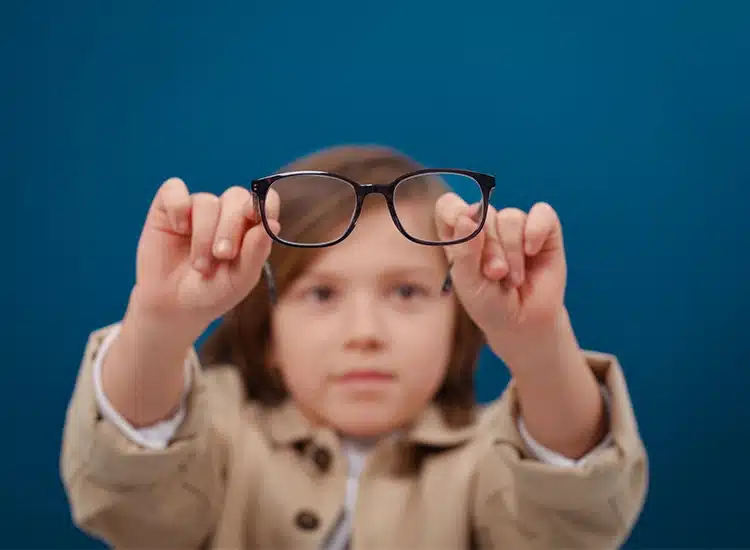 Child holding a pair of black eyeglasses with both hands in front of their face, featuring a high-quality Rodenstock eyeglass lens, standing against a plain blue background.