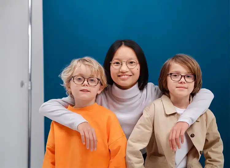 Three children wearing Rodenstock eyeglass lenses stand in front of a blue background. The child in the center has arms around the other two, and all three are looking at the camera.