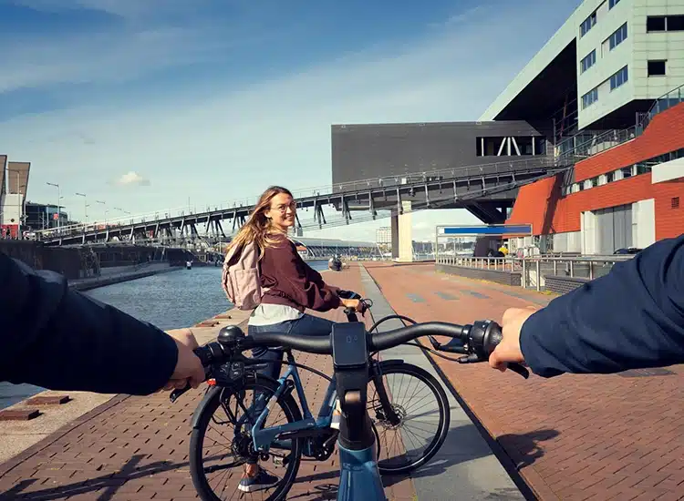 Two people ride bicycles along a canal-side path in an urban area; one person looks back and smiles, enjoying the vibrant cityscape through their Rodenstock Lens, while modern buildings are visible in the background.