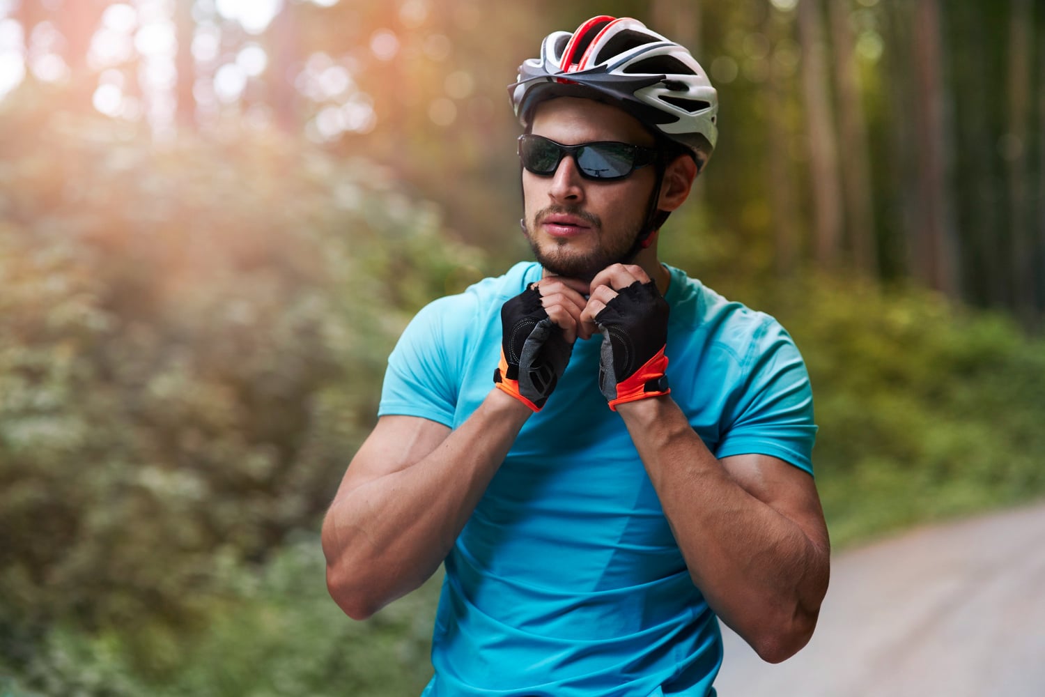 A man in a blue shirt, helmet, and sunglasses with Rodenstock lenses fastens his bike helmet strap while standing on a wooded path.