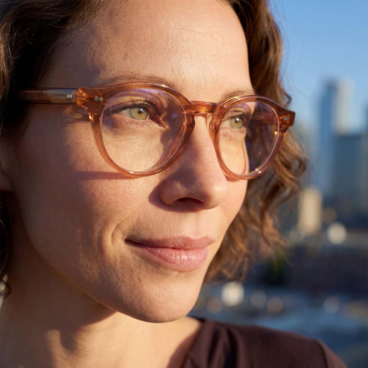 A woman with light brown curly hair and clear-framed glasses looks into the distance outdoors, with a blurred cityscape in the background.