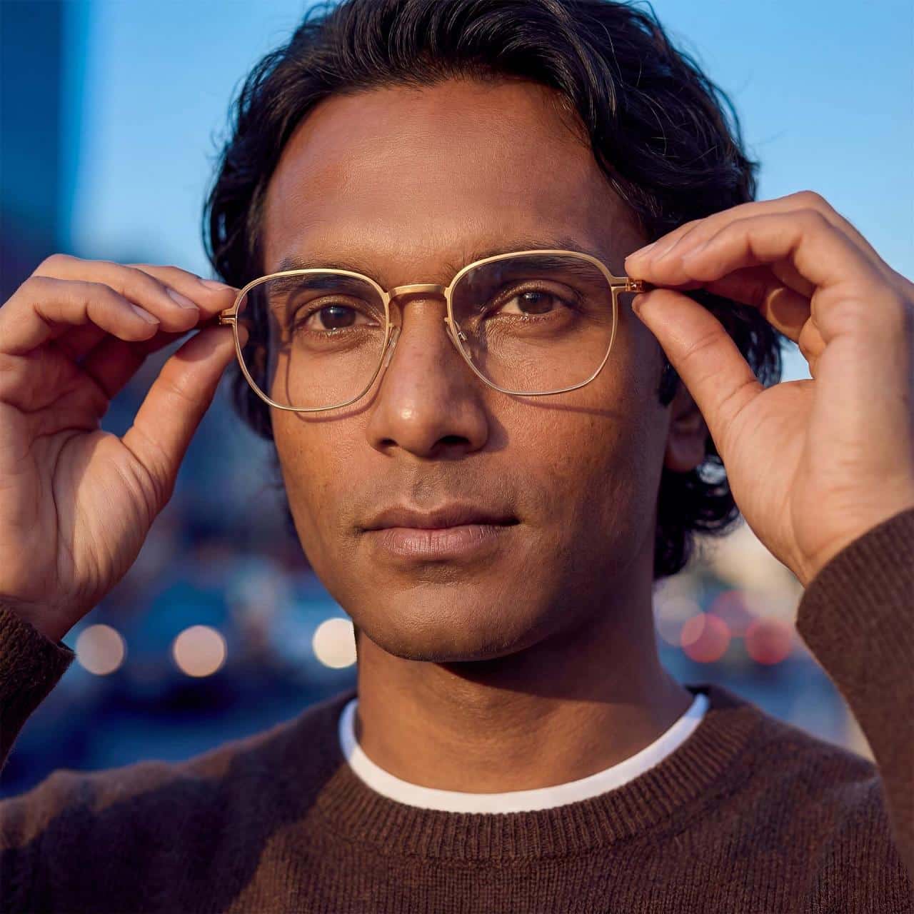 A person with medium skin tone and dark hair adjusts their gold-rimmed glasses outdoors, wearing a brown sweater and white undershirt.