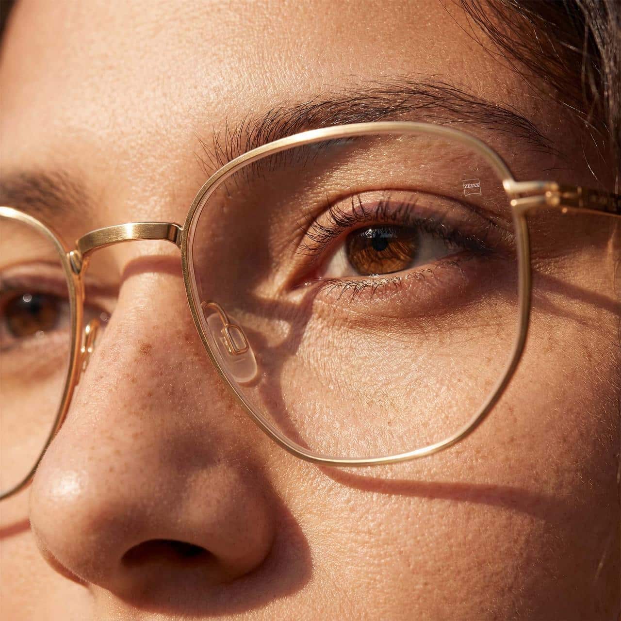 Close-up of a person’s face showing brown eyes, freckles, and gold-rimmed eyeglasses reflecting light.