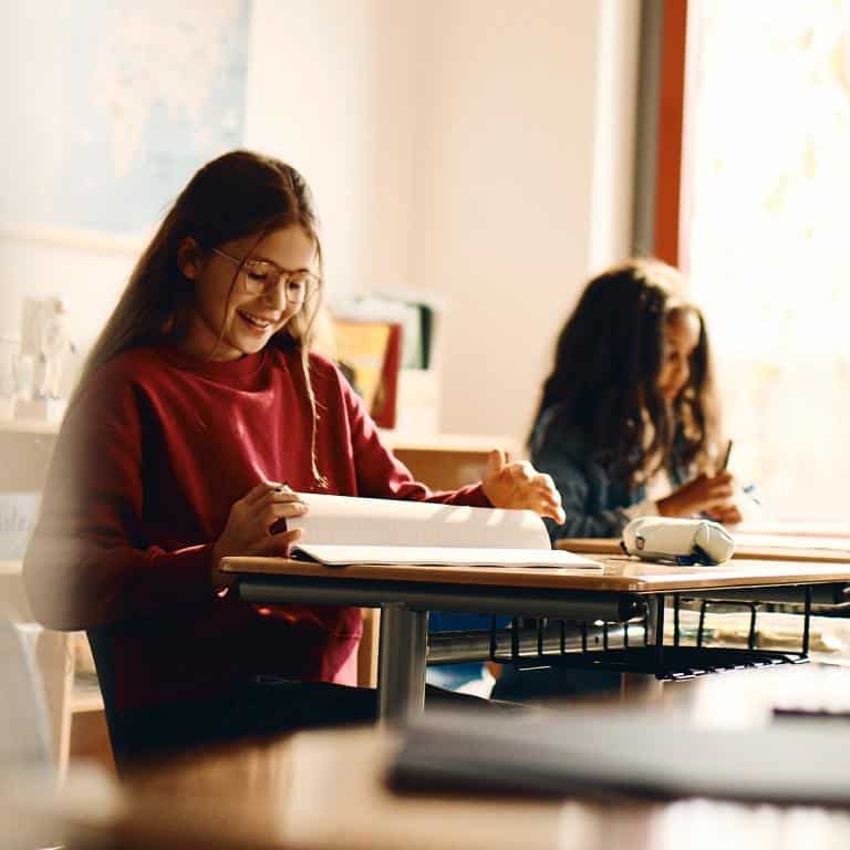 Two girls sit at desks in a classroom, one in the foreground smiling and looking at a notebook, the other in the background writing.
