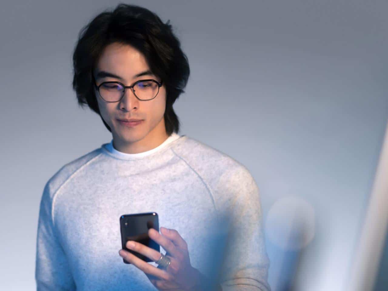 A person wearing glasses and a light gray sweatshirt looks at a smartphone in their hand against a plain background.