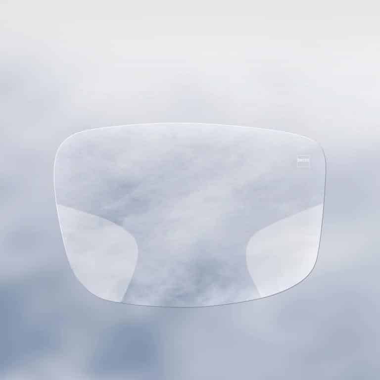 A single, transparent eyeglass lens with faint reflections, displayed against a soft, cloudy background.
