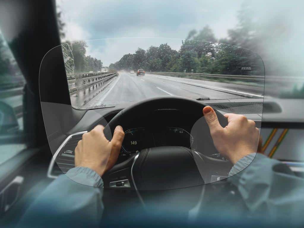 View from inside a car showing the driver’s hands on the wheel, looking through a clear lens on a rainy road with blurred surroundings.