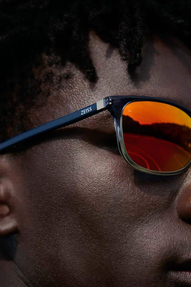 Close-up of a person wearing Zeiss sunglasses, reflecting a sunset and a road in the orange-tinted lenses.