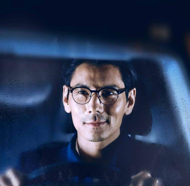 A man with glasses sits in a car, viewed through a windshield with condensation or raindrops, looking forward with a neutral expression.
