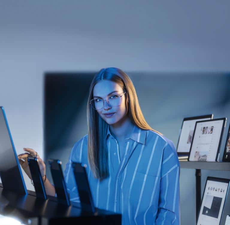 A woman with long hair and glasses sits at a desk with monitors and framed pictures, wearing a blue striped shirt, in a modern office setting.
