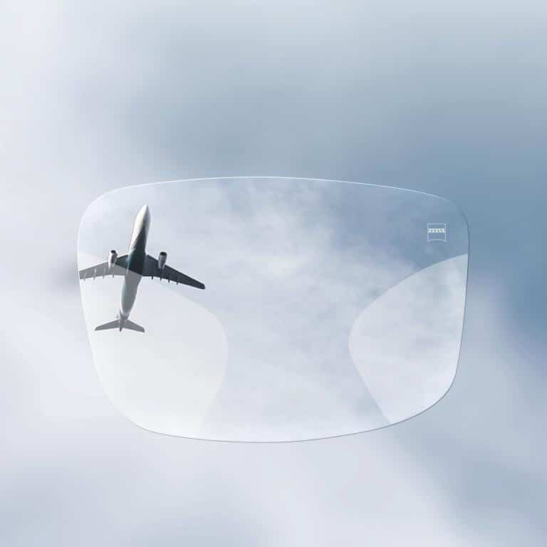 A commercial airplane flies overhead, viewed through a single eyeglass lens floating against a cloudy sky.