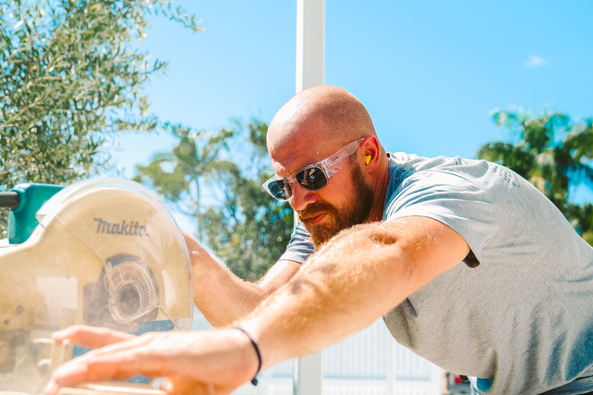 A man wearing protective glasses and earplugs operates a Makita miter saw.